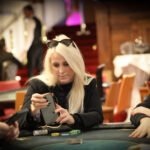 Ein Mann in einem eleganten Anzug sitzt an einem Pokertisch und hält zwei Spielkarten in der Hand. Er schaut konzentriert auf seine Karten, während im Hintergrund andere Spieler und Pokerchips zu
