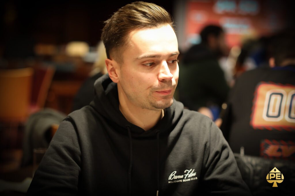 Ein Mann sitzt an einem Pokertisch und studiert konzentriert seine Karten. Um ihn herum sind Chips und Spielkarten verstreut. Im Hintergrund sind unscharfe Personen zu sehen, die ebenfalls Poker spielen