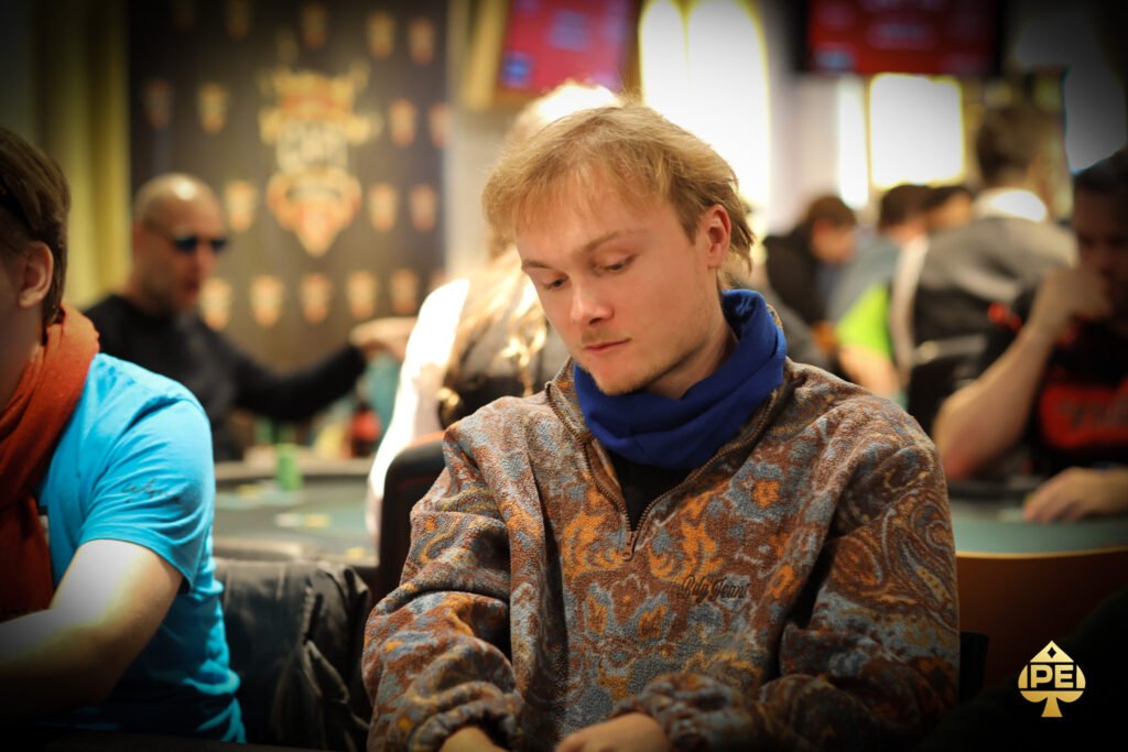 Ein Mann sitzt an einem Pokertisch mit grünem Filz und hält seine Karten in der Hand. Er trägt eine dunkle Sonnenbrille und konzentriert sich auf das Spiel. Im Hintergrund sind