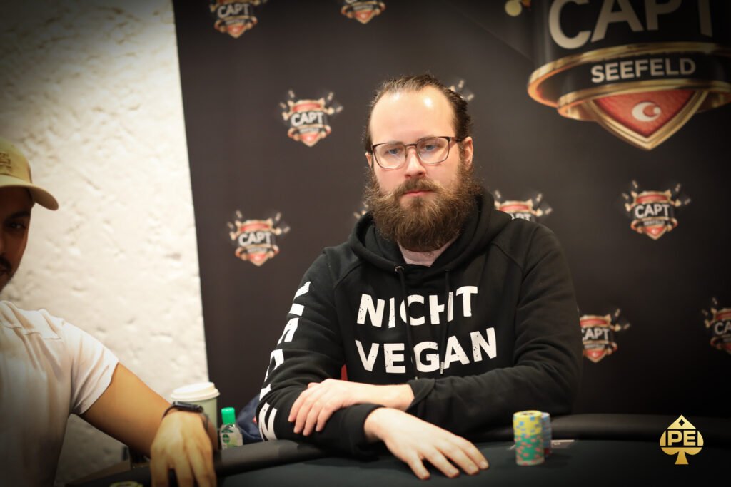 Ein Mann in einem schwarzen Anzug sitzt an einem Pokertisch und hält in der Hand ein Ass und eine Zwei in Herz. Er wirkt konzentriert und umgeben von anderen Pokerspielern in einem eleganten