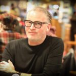 Ein Mann sitzt an einem Pokertisch und hält zwei Spielkarten in der Hand, während er konzentriert auf seine Gegner schaut. Im Hintergrund sind Pokerchips und Getränke zu sehen.