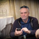 Ein Mann mit einem roten Hemd und einer schwarzen Weste sitzt an einem Pokertisch und hält seine Karten in der Hand. In der Mitte des Tisches liegen Chips und Spielkarten. Im Hintergrund