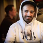 Ein Mann in einem schwarzen Anzug und einer Krawatte sitzt an einem Pokertisch mit Pokerchips und Karten. Er hat eine ernste Miene und schaut konzentriert auf seine Karten. Im Hinter