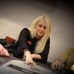 Ein Mann in einem schwarzen Anzug und einer Krawatte sitzt an einem Pokertisch und hält seine Karten in der Hand, während er konzentriert auf die Mitte des Tisches schaut. Um ihn herum