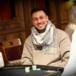 Ein Mann in einem schwarzen Anzug und einer schwarzen Sonnenbrille spielt Poker an einem eleganten Tisch in einem luxuriösen Casino. Er hält Karten in der Hand und hat einen ernsten Gesichtsaus
