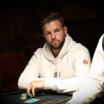 Ein Mann in einem schwarzen Anzug sitzt an einem Pokertisch mit grünem Filz und hält zwei Spielkarten in der Hand. In der Mitte des Tisches liegen weitere Karten und Pokerchips. Im