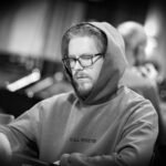 Ein Mann sitzt an einem Pokertisch und hält zwei Karten in der Hand, während er konzentriert auf die Gemeinschaftskarten schaut. Um ihn herum sind Pokerchips und ein Stapel Karten zu