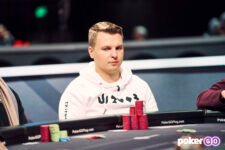 Ein Mann sitzt an einem Pokertisch während eines Turniers der World Series of Poker. Er hat eine Sonnenbrille auf und betrachtet konzentriert seine Karten. Im Hintergrund sind andere Spieler und Dealer zu sehen