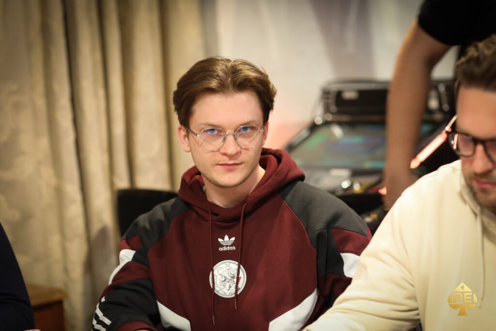 Ein Mann mit dunklen Haaren und einem dunklen Anzug sitzt lächelnd an einem Pokertisch und hält Karten in der Hand.