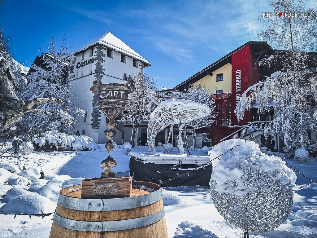 Ein Mann in winterlicher Kleidung und mit einer Mütze steht neben einem Schneemann in einem verschneiten Park. Im Hintergrund sind Bäume mit Schnee bedeckt.