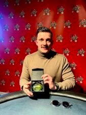 Ein Mann mit schwarzer Sonnenbrille und Kappe sitzt an einem Pokertisch und studiert konzentriert seine Karten. In der Hintergrund sind weitere Spieler und Pokerchips zu sehen.