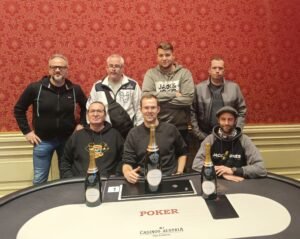 Ein Mann in einem schwarzen Anzug und einer roten Krawatte sitzt an einem Pokertisch und hält Pokerkarten in der Hand. Im Hintergrund sind andere Spieler und Chips zu sehen.