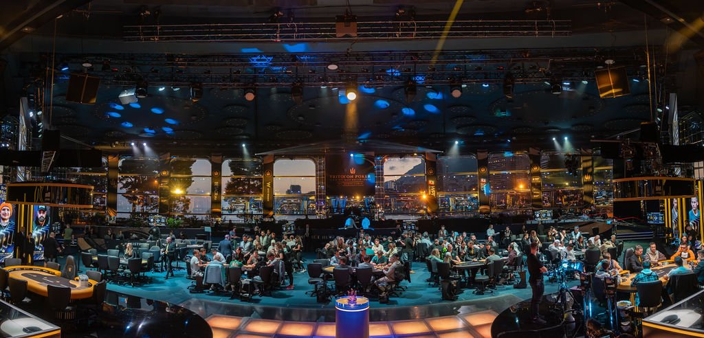 Ein Panoramablick auf einen großen Pokerraum voller Spieler, die an Tischen sitzen und Pokerturniere spielen. Die Spieler sind konzentriert und die Atmosphäre ist gespannt.