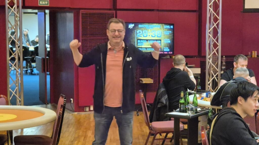 Ein Mann in einem schwarzen Anzug und einer roten Krawatte sitzt an einem Pokertisch und hält zwei Könige (King of Hearts und King of Diamonds) in der Hand. Er hat einen ernsten Gesichtsa