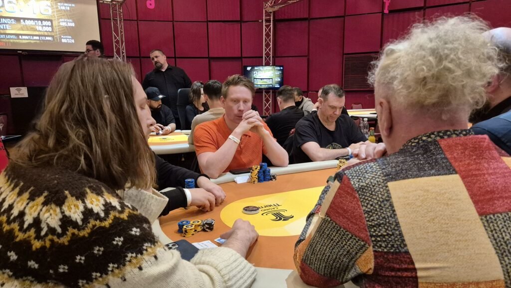Ein professioneller Pokertisch mit grüner Filzoberfläche, umgeben von gepolsterten Stühlen. Auf dem Tisch liegen Spielkarten, Pokerchips und Getränke. Im Hintergrund