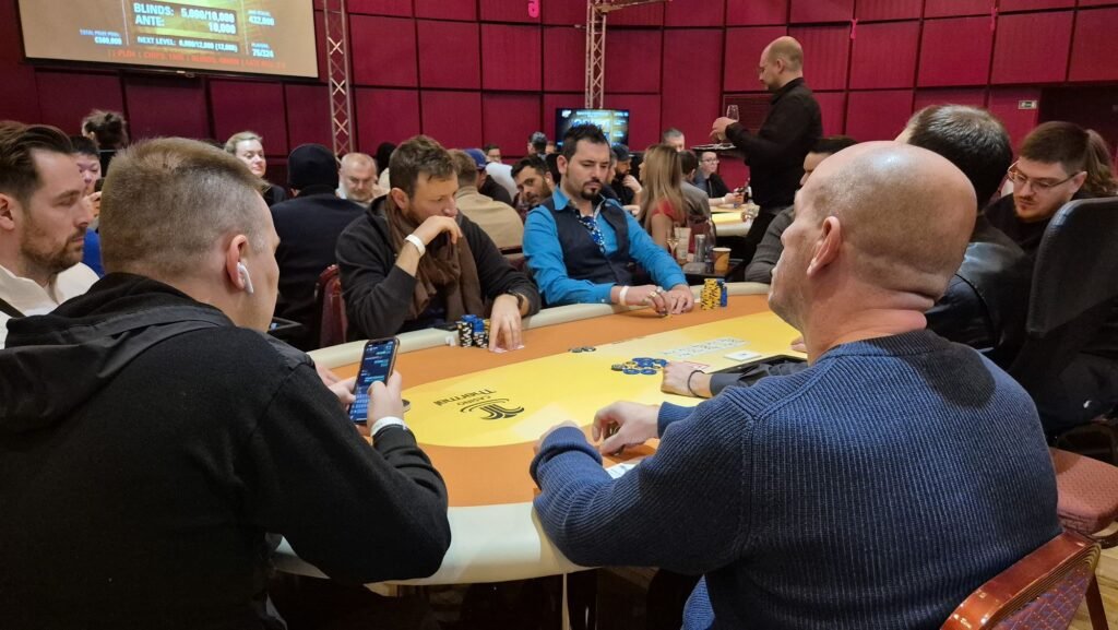 Ein Mann sitzt an einem Pokertisch und hält zwei Karten in der Hand. Er trägt eine dunkle Brille und konzentriert sich auf das Spiel. Um ihn herum liegen Pokerchips und Karten auf
