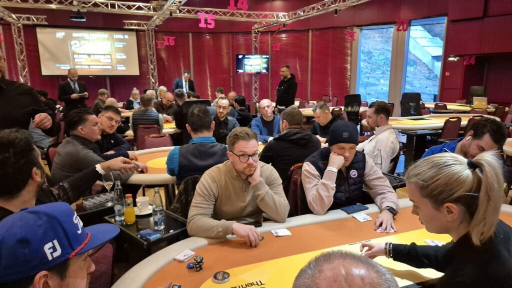 Ein Pokertisch mit grüner Filzdecke, umgeben von Spielkarten, Pokerchips und Getränken. Vier Personen sitzen um den Tisch herum, konzentriert auf ihre Karten.