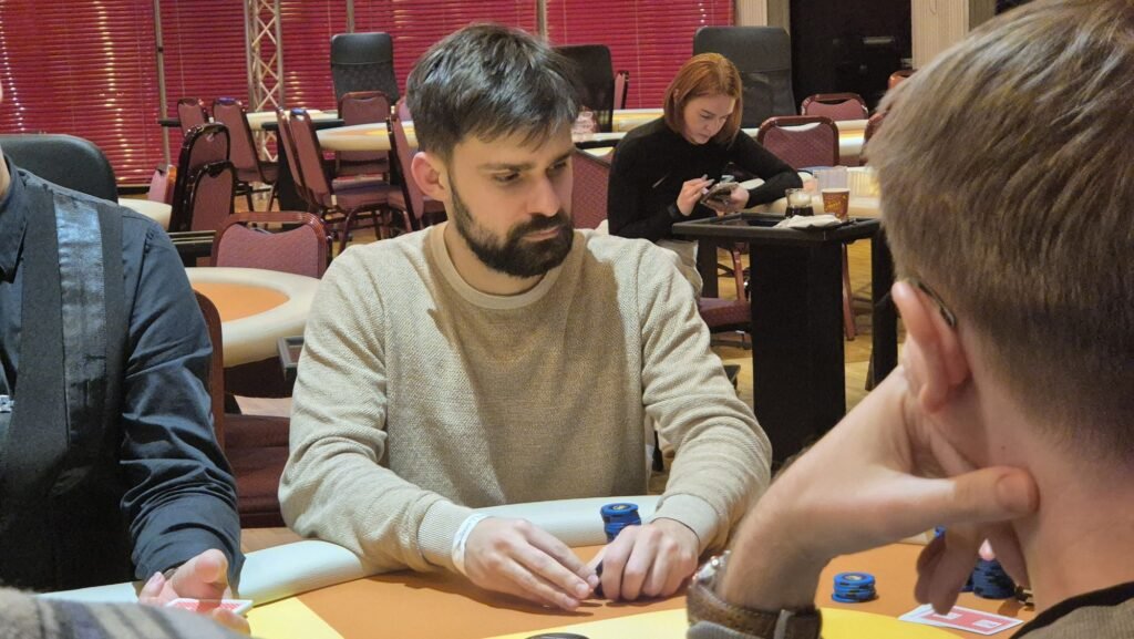 Ein professioneller Pokertisch mit grünem Filz und einem Kartendeck in der Mitte, umgeben von Pokerchips und Getränken. Im Hintergrund sind Spieler und Dealer zu sehen, die Karten halten und