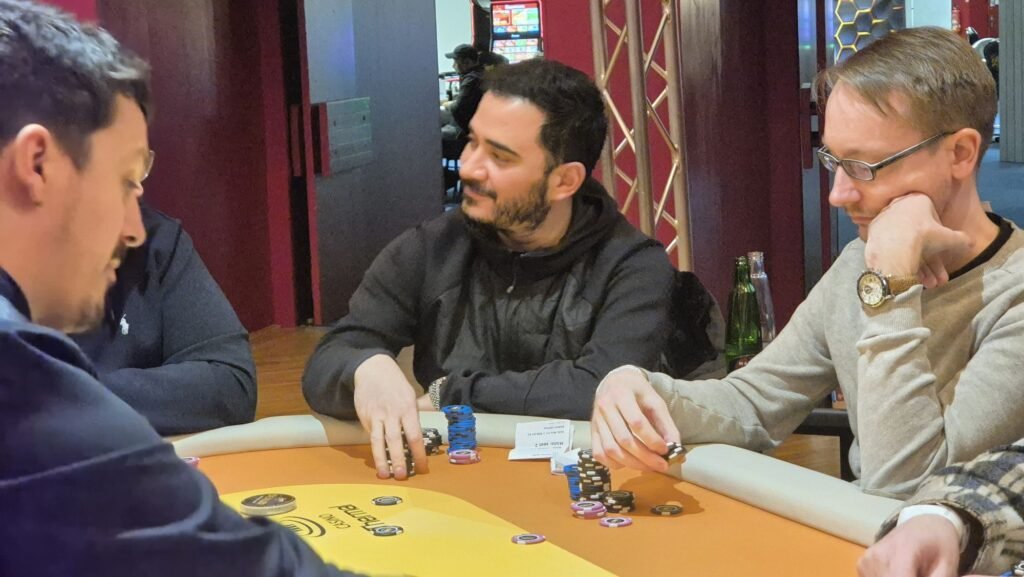Ein Tisch mit einem grünen Pokertuch, auf dem Spielkarten und Pokerchips liegen. In der Mitte des Tisches befindet sich ein Stapel von Jetons. Im Hintergrund sind Spielerhände zu