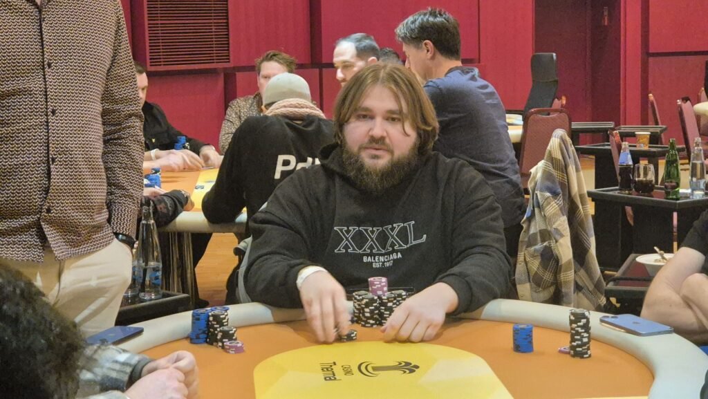 Ein Mann in einem dunkelblauen Anzug sitzt an einem Pokertisch mit grünem Filz. Er hält seine Spielkarten in der Hand und schaut konzentriert auf den Tisch. Im Hintergrund