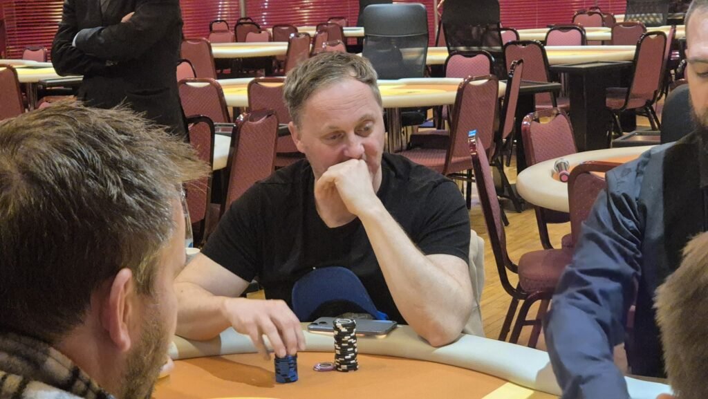 Ein Mann in einem dunklen Anzug und einer Krawatte sitzt an einem Pokertisch und hält zwei Spielkarten in der Hand. In der Mitte des Tisches liegen weitere Karten. Im Hintergrund sind andere