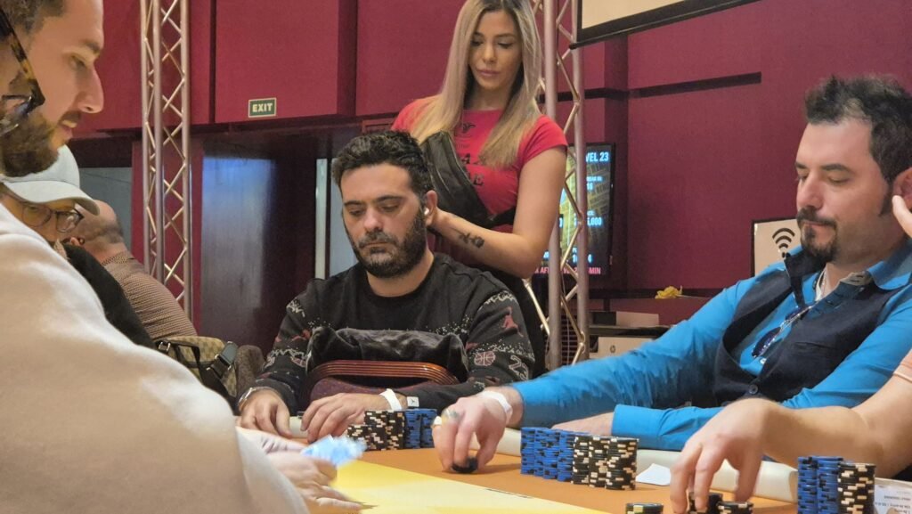 Ein Mann spielt Poker an einem Tisch mit grüner Filzdecke. Er hat eine Hand mit König-Karten. Andere Spieler und Pokerchips sind im Hintergrund zu sehen.