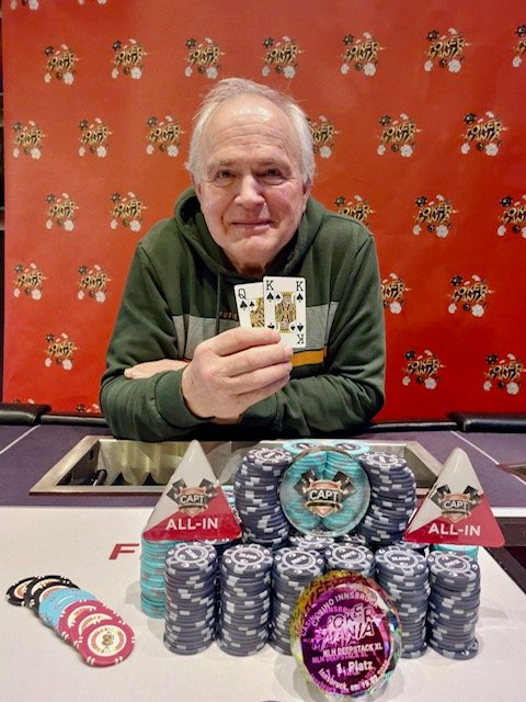 Ein Mann und eine Frau sitzen lächelnd an einem Pokertisch, während sie Karten halten. Die Frau hat rote Haare und trägt eine schwarze Bluse, der Mann hat dunkle Haare und trägt ein