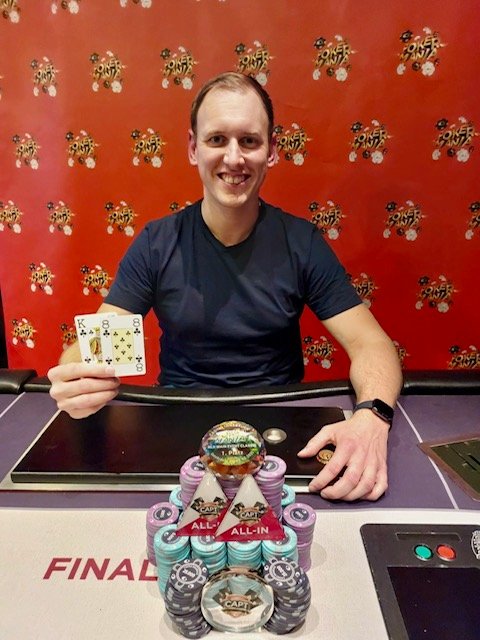 Ein Mann und eine Frau sitzen an einem Pokertisch. Der Mann hält Karten in der Hand, während die Frau konzentriert auf ihre Karten schaut. Im Hintergrund sind Pokerchips und Getränke