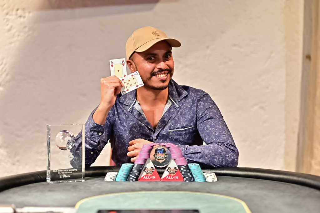Ein Mann in einem schwarzen Anzug sitzt an einem Pokertisch und hält Karten in der Hand, während er konzentriert auf das Spielgeschehen schaut. Im Hintergrund sind weitere Pokerspieler zu
