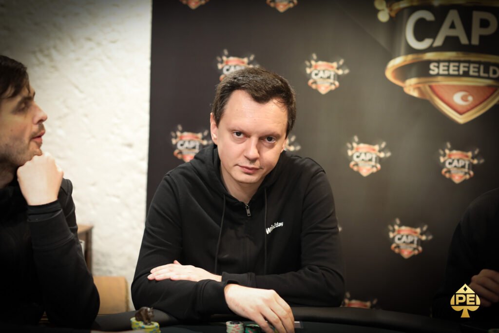 Ein Mann sitzt an einem Pokertisch mit Chips und Karten vor sich. Er hat eine ernste Miene und trägt ein dunkles Hemd.