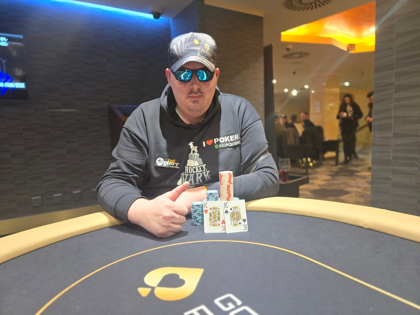 Ein Foto von einem Pokertisch während eines Turniers mit Stapeln von Pokerchips und Spielkarten darauf. Die Spieler sind konzentriert und entschlossen, während sie ihre Hände spielen. Ein Dealer sitzt am Tisch