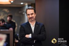 Ein Mann mit dunklen Haaren und Brille, der konzentriert an einem Pokertisch sitzt und Karten betrachtet.