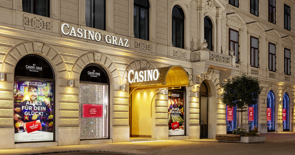 Ein nächtliches Foto des Casino Graz mit beleuchteter Fassade und dem Eingangsbereich. Das Gebäude strahlt in verschiedenen Farben und spiegelt sich in der Dunkelheit wider. Menschen sind vor