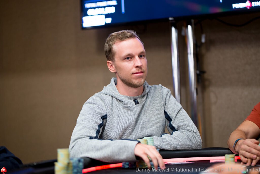 Ein junger männlicher Pokerspieler konzentriert sich am Tisch während eines Turniers. Er hat kurze dunkle Haare, trägt ein schwarzes Hemd und hat eine Sonnenbrille auf der Stirn.