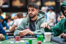 Ein Mann in einem dunkelblauen Anzug und weißem Hemd, Fabian Bernhauser, sitzt an einem Pokertisch und betrachtet konzentriert seine Karten. Im Hintergrund sind andere Spieler und Poker