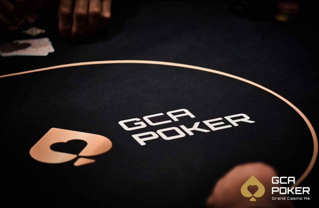 Ein Mann sitzt an einem Pokertisch und hält zwei Spielkarten in der Hand, während er konzentriert auf seine Gegner schaut. Die anderen Spieler sind nicht im Bild zu sehen.