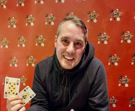 Ein Mann spielt Poker an einem Tisch in einem eleganten Casino. Er hat einen ernsten Gesichtsausdruck und hält Karten in der Hand, während er von anderen Spielern umgeben ist. Die Szene strah