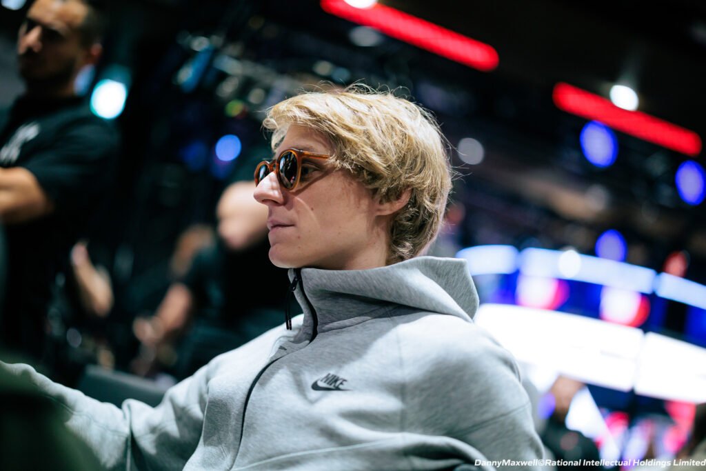 Ein junger Mann namens Lukas Hafner sitzt an einem Pokertisch und sieht konzentriert auf seine Karten. Er hat kurze dunkle Haare und trägt ein dunkles Hemd. Im Hintergrund