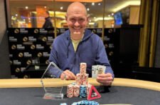 Ein Mann mit dunklem Haar und Bart, der eine Sonnenbrille trägt und eine Pokerkarte in der Hand hält, während er konzentriert in die Kamera schaut.