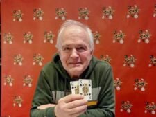 Ein Mann mit dunklen Haaren und einem gestreiften Hemd sitzt an einem Pokertisch und hält Karten in der Hand. Er schaut konzentriert auf seine Karten.