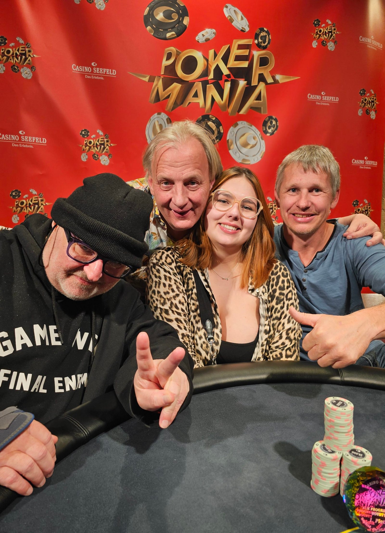 Ein Mann in einem schwarzen Anzug und einer schwarzen Sonnenbrille sitzt an einem Pokertisch und hält zwei Spielkarten in der Hand, während er konzentriert auf seine Gegner schaut.