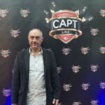 Ein Mann sitzt an einem Pokertisch und hält zwei Spielkarten in der Hand, während er konzentriert auf seine Gegner schaut. Im Hintergrund sind weitere Spieler und Chips auf dem Tisch zu sehen.