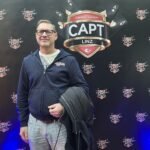 Ein Tisch mit einem Pokerspiel, bei dem vier Personen Karten halten und Pokerchips stapeln. In der Mitte liegen Spielkarten und weitere Chips. Die Spieler sind konzentriert und haben ernste Gesichtsa