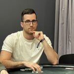 Ein Mann und eine Frau sitzen an einem Pokertisch und spielen Karten. Der Mann hat eine Hand mit Pokerchips vor sich, während die Frau ihre Karten betrachtet. Im Hintergrund sind weitere Personen zu sehen, die