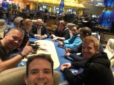 Ein Gruppenbild von Pokerspielern, die an einem Cash Game-Tisch im King's Casino spielen. Die Spieler sitzen um einen grünen Pokertisch herum und halten ihre Karten in der Hand, während sie konzent