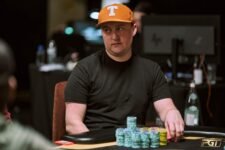 Ein Mann in einem dunklen Anzug sitzt an einem Pokertisch mit Chips und Karten vor sich. Im Hintergrund sind weitere Spieler zu sehen.