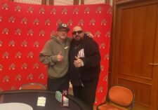 Ein Mann mit einer Sonnenbrille und einem Hut sitzt an einem Pokertisch und hält zwei Asse als Startkarten. Die anderen Spieler am Tisch sind verschwommen im Hintergrund zu sehen.