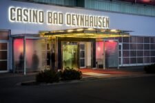 Ein Foto des Außenbereichs des Casinos Bad Oeynhausen, das von Bäumen umgeben ist und eine elegante Architektur zeigt. In der Mitte des Bildes ist der Eingang des Casinos mit großen