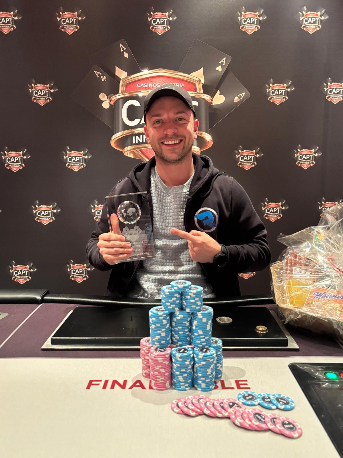 Ein Mann in einem eleganten blauen Anzug und einer schwarzen Sonnenbrille spielt Poker an einem Tisch in einem luxuriösen Casino. Er hat eine ernste Miene und hält eine Hand mit Spielkarten.