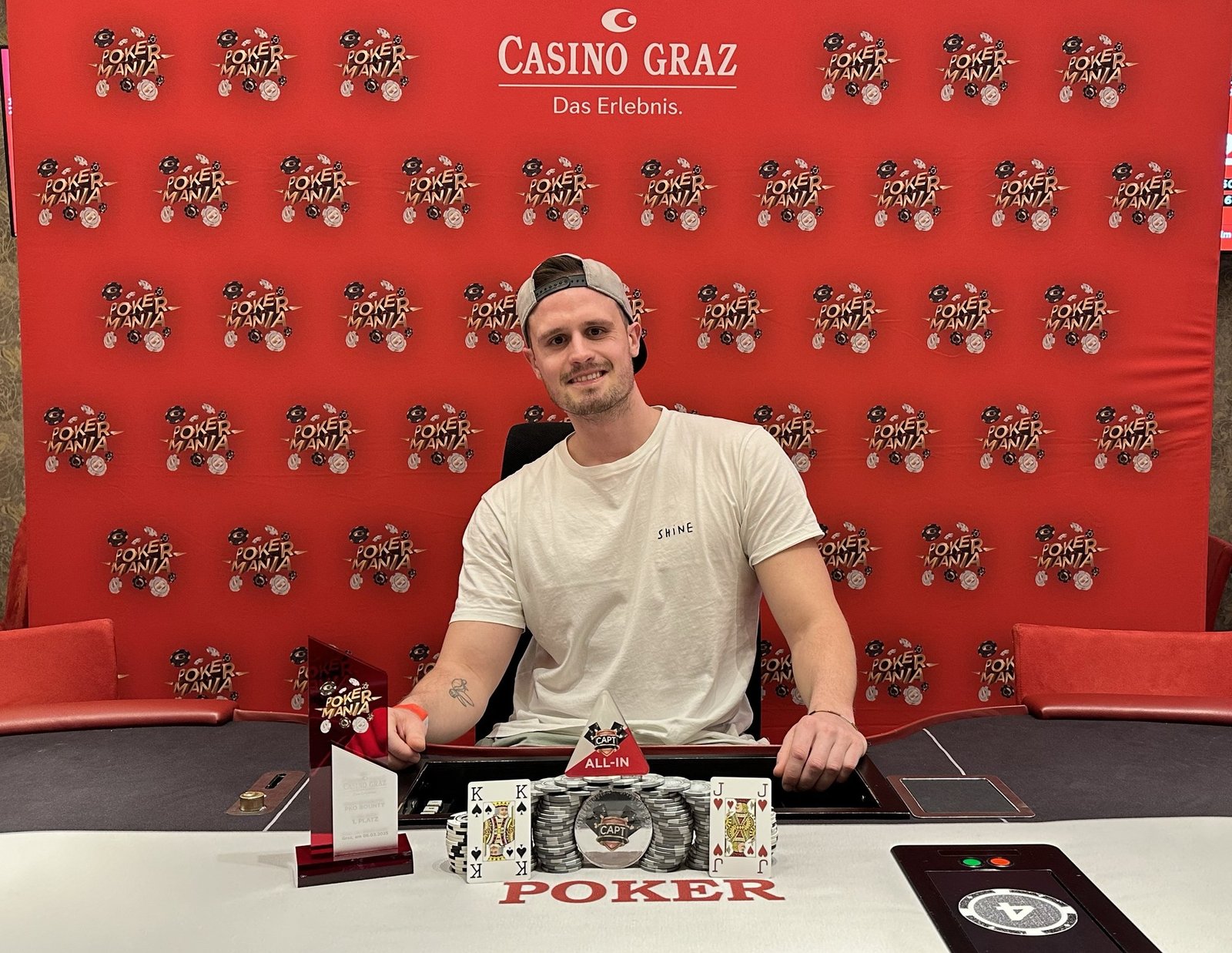 Ein Mann freut sich über seinen Sieg beim Pokerspiel. Er hält stolz seine Karten in der Hand und lächelt glücklich. Um ihn herum stehen andere Spieler, die enttäuscht wirken